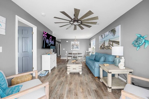 A living room with a ceiling fan, a blue sofa, a dining table, and a white cabinet. - Totally Beachin' - Oceanfront, King Bed, Pool (North Myrtle Beach)