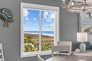 A room with a large window overlooking a beach, a chandelier, and a table with a lamp. - Totally Beachin' - Oceanfront, King Bed, Pool (North Myrtle Beach)
