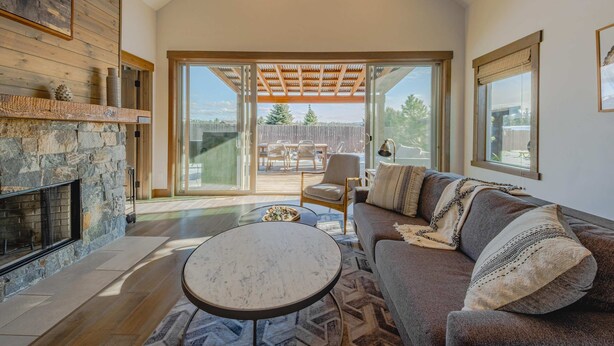 Un salon spacieux doté d’une cheminée en pierre, d’un grand canapé, d’une table basse ronde et offrant une vue sur une terrasse extérieure, accessible par une porte-fenêtre coulissante.