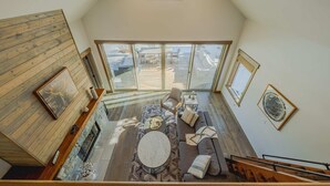 Un espace de vie de style loft avec un plafond haut, des poutres en bois, une cheminée, un ensemble de canapés et une vue sur une terrasse extérieure, accessible par de grandes portes vitrées.