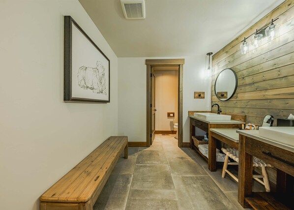 Un baño con un banco de madera, una obra de arte enmarco, un espejo y dos lavamanos.