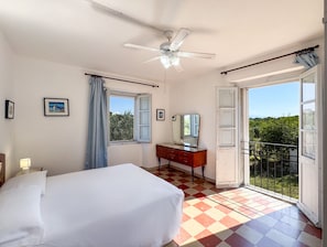 A bedroom with a bed, a dresser, a ceiling fan, a window with curtains, and a balcony with a view.