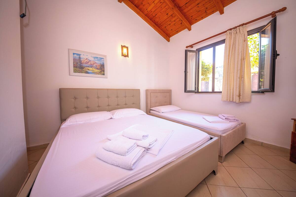 A room with two beds, a painting on the wall, a window with curtains, and a wooden ceiling.