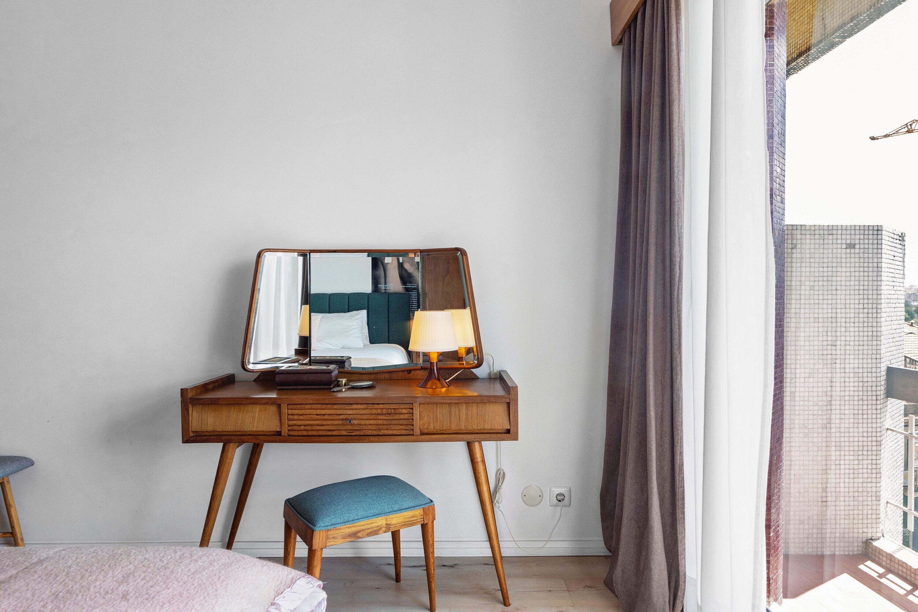 A hotel room with a wooden desk, a mirror, a lamp, a bed with a blue cushion, and a view of a building through a window.