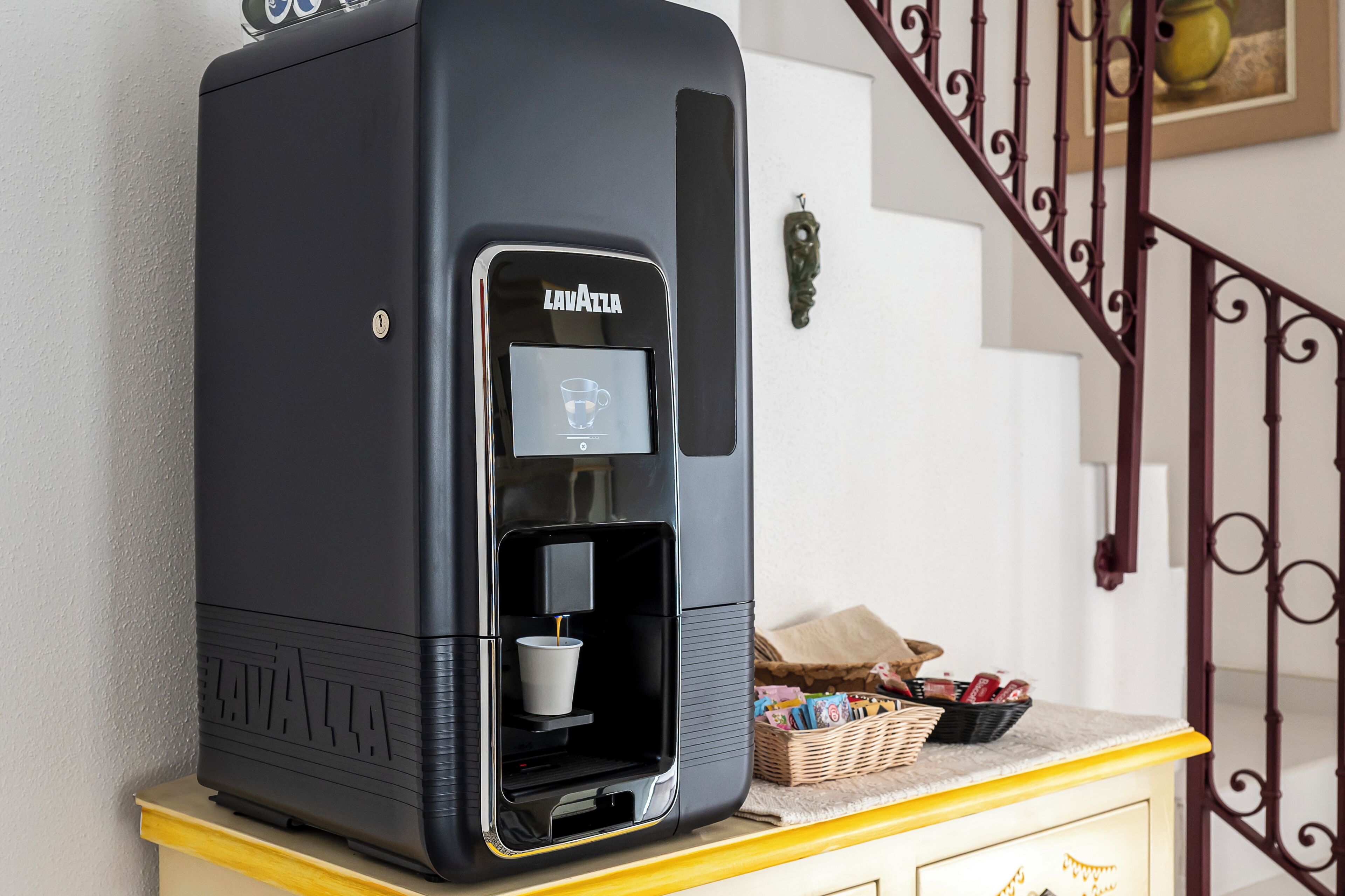 A coffee machine with a cup dispenser, a basket with snacks, and a staircase in the background.