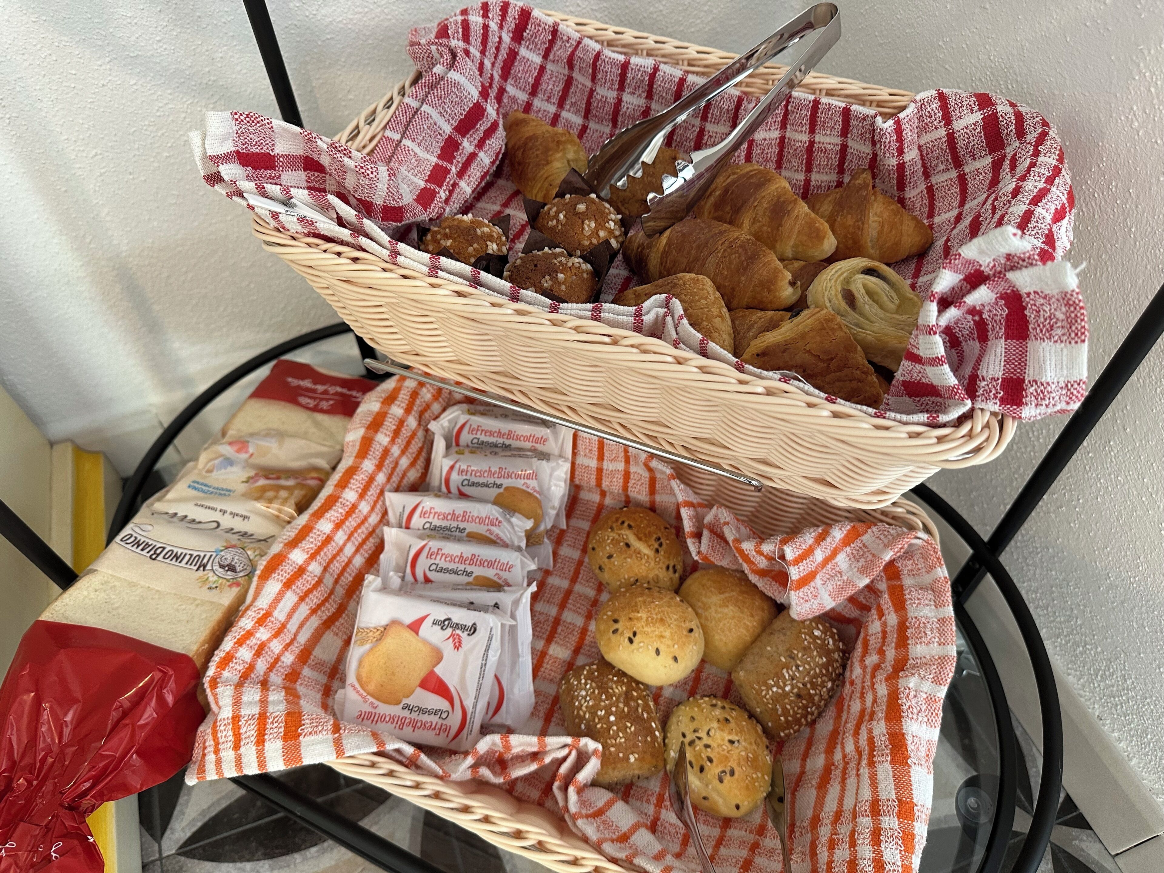 Una cesta de mimbre llena de productos horneados, como croissants y bollos, sobre una mesa con mantel a cuadros.