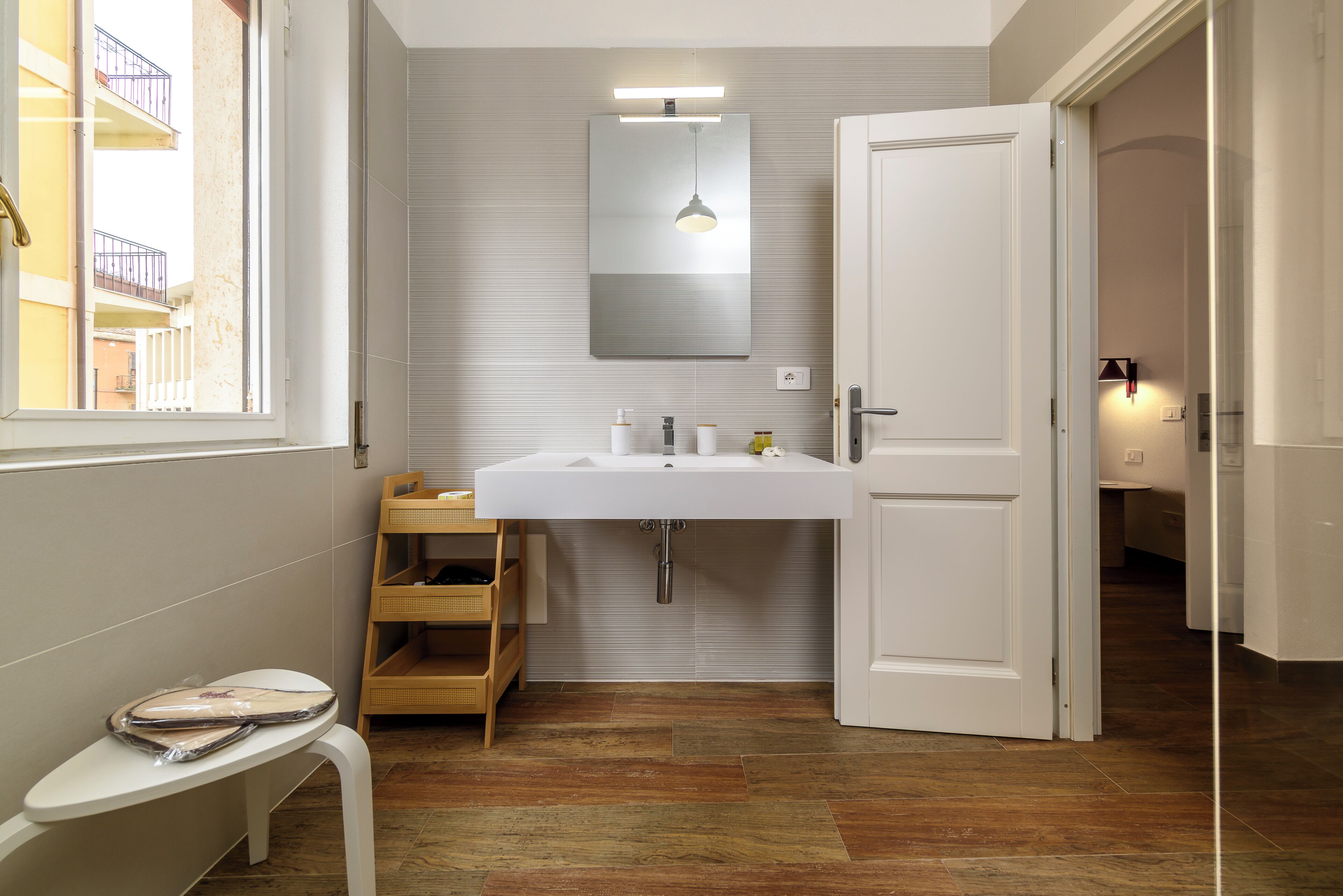 Une salle de bain avec un lavabo blanc, un miroir et un plancher en bois. Il y a une fenêtre donnant sur un bâtiment à l’extérieur.
