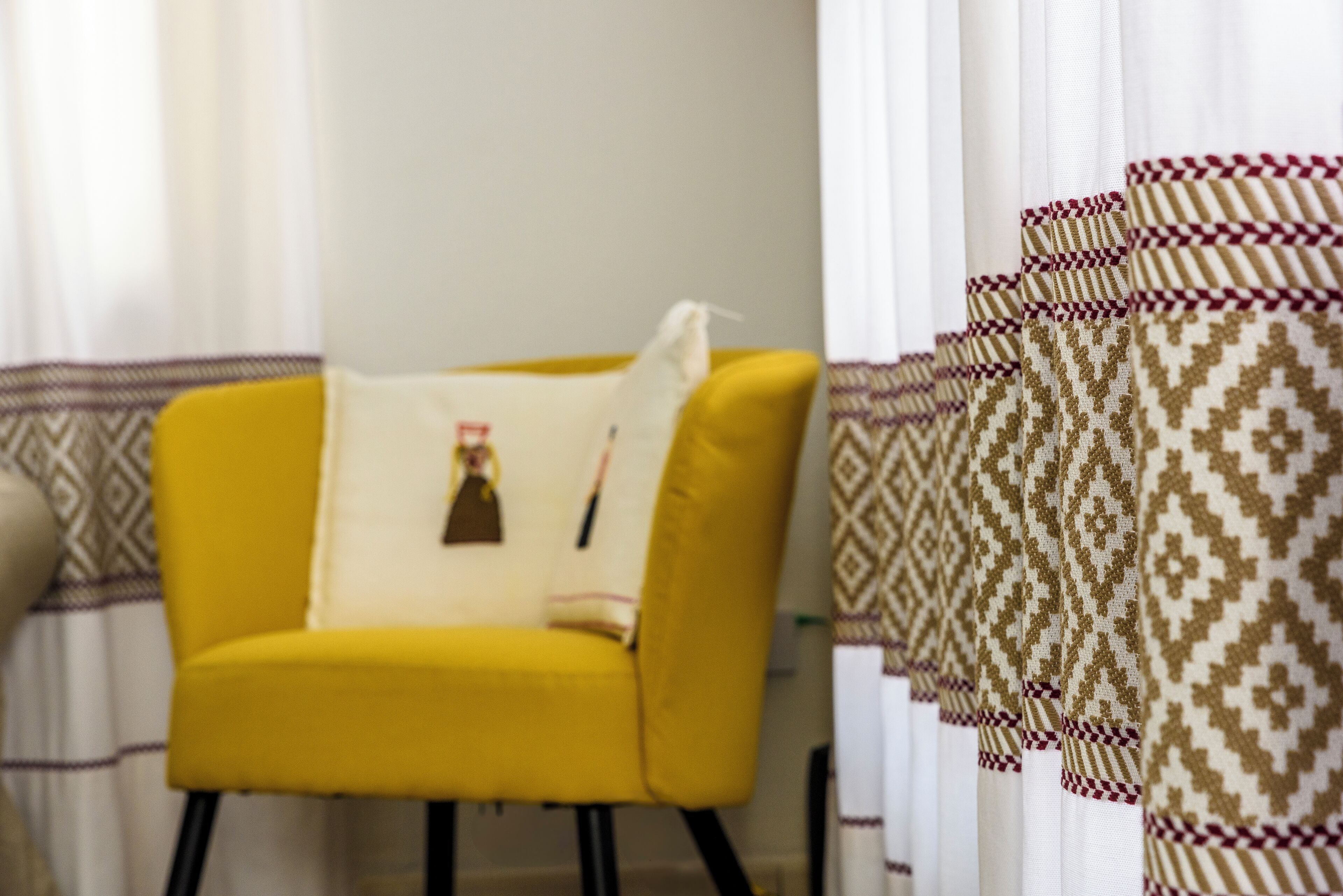 Un fauteuil jaune avec un coussin, placé près d’une fenêtre ornée de rideaux à motifs.
