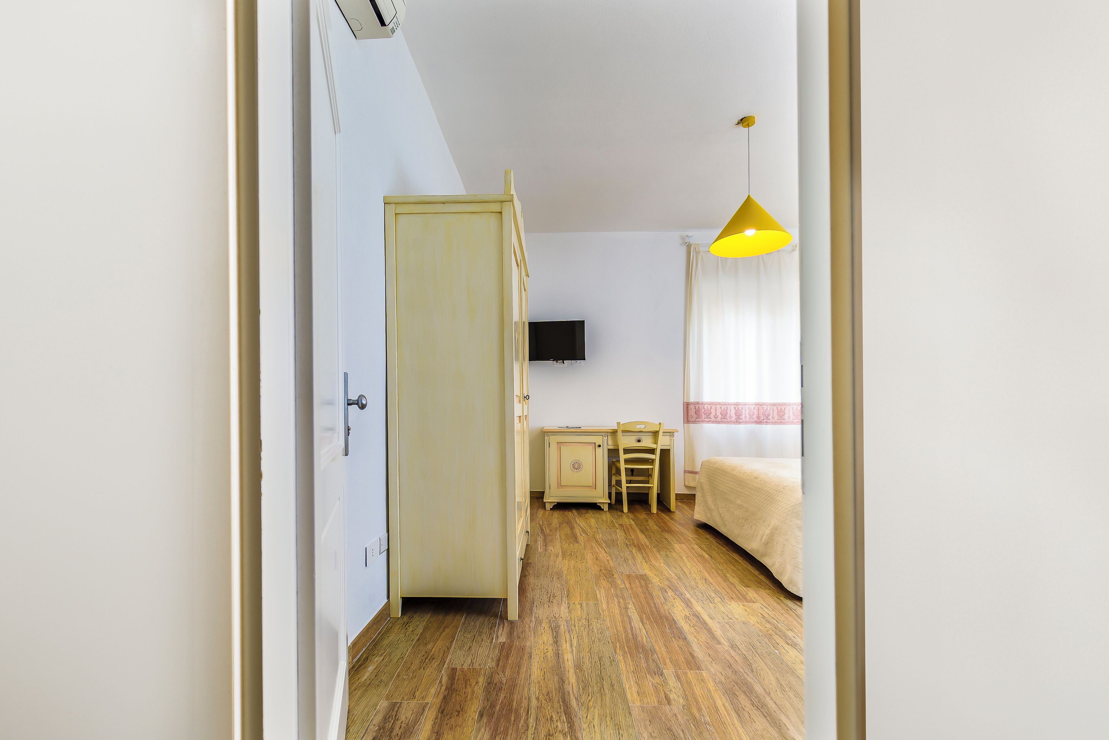 A hotel room with a wooden floor, a bed, a desk, a chair, a television, and a yellow pendant light.