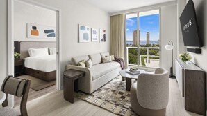 A modern hotel room with a bed, sofa, armchair, and a large window offering a city view. - Modern 1BR Retreat in Miami – Bay Views (Miami)