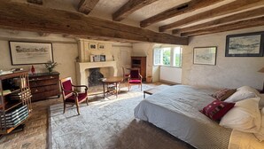 Une pièce rustique avec des poutres apparentes, une cheminée, un lit recouvert d’une courtepointe à rayures et un bureau avec des livres.