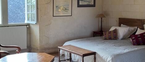 Un dormitorio con techo de madera, una cama con ropa de cama a rayas, una mesita de noche, una lámpara y una ventana con vista a un muro de ladrillos.
