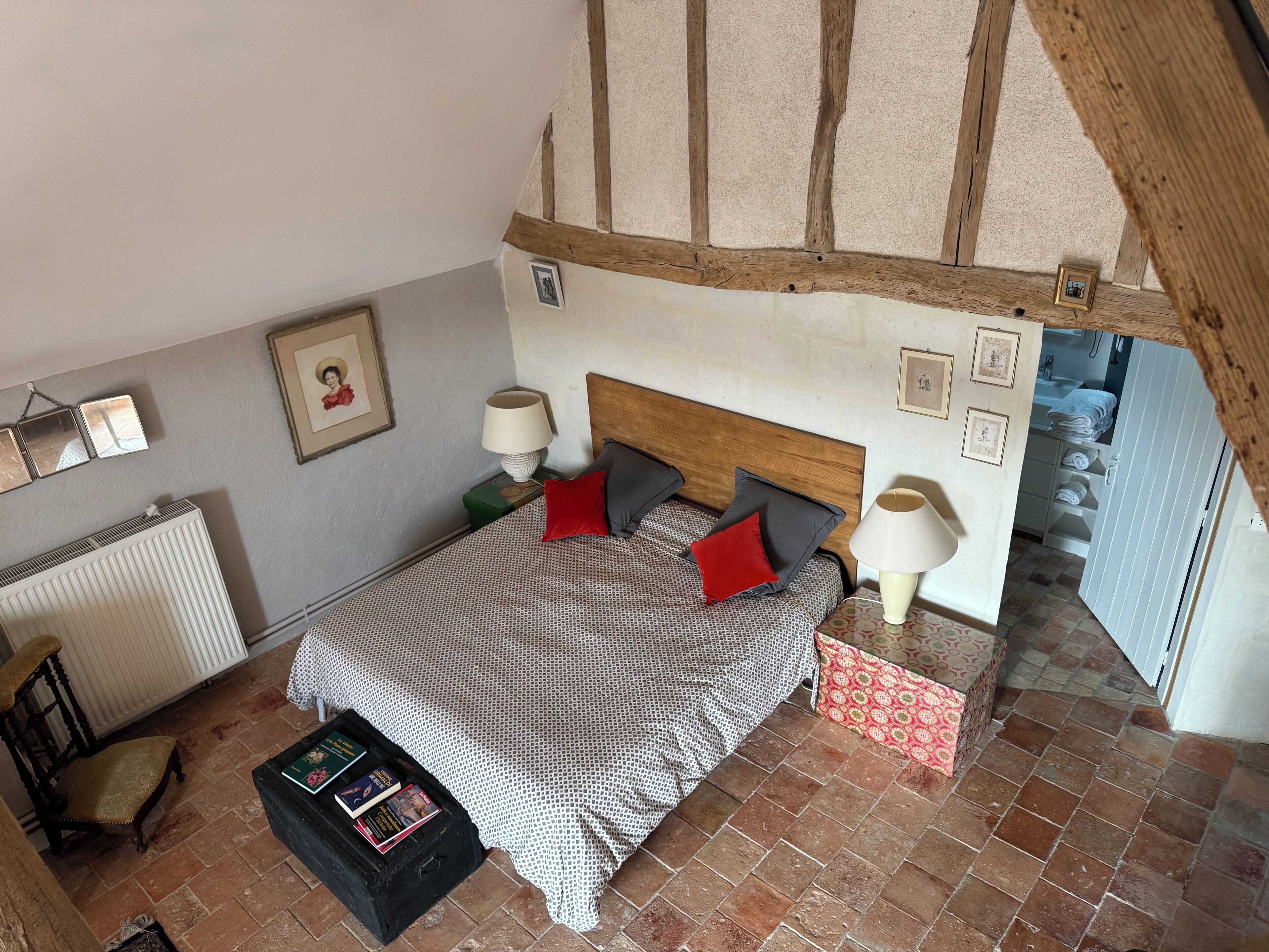 A bedroom with a wooden ceiling, a bed with patterned bedding, a bedside table with a lamp, a radiator, a chair, and a small table with books.