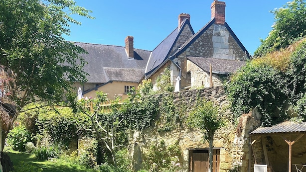 Un cadre extérieur rustique avec un bain à remous en bois, un mur de pierre et une maison traditionnelle en arrière-plan.