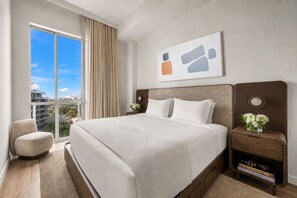 Een hotelkamer met een groot bed, uitzicht op de stad, een fauteuil en een nachtkastje met een bloevas.