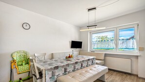 A dining area with a table, chairs, a bench, a child’s high chair, a clock, a TV, and a window with a view.