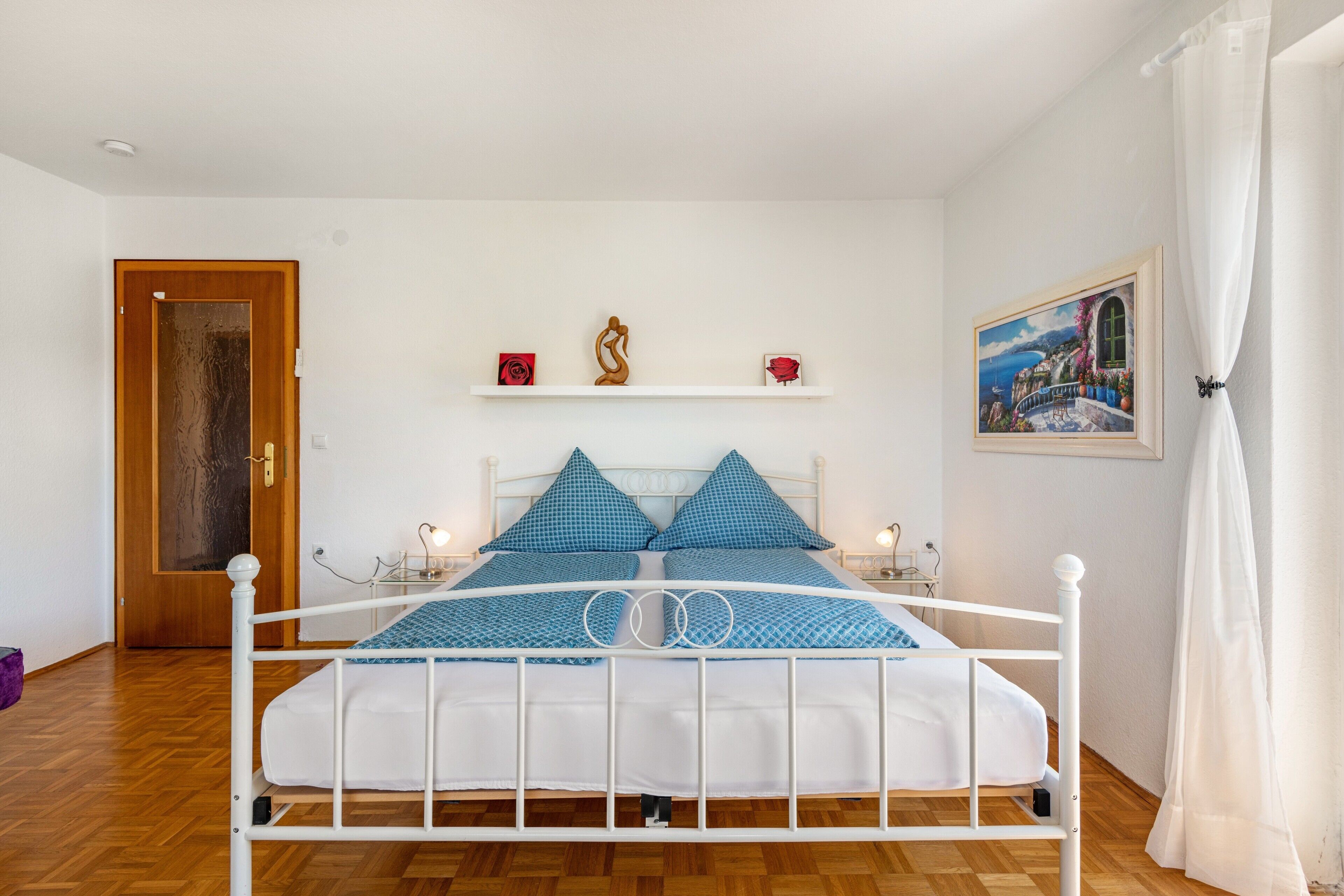 Un dormitorio con estructura de cama de metal blanco, ropa de cama azul, una puerta de madera, un estante blanco con objetos decorativos y un cuadro en la pared.