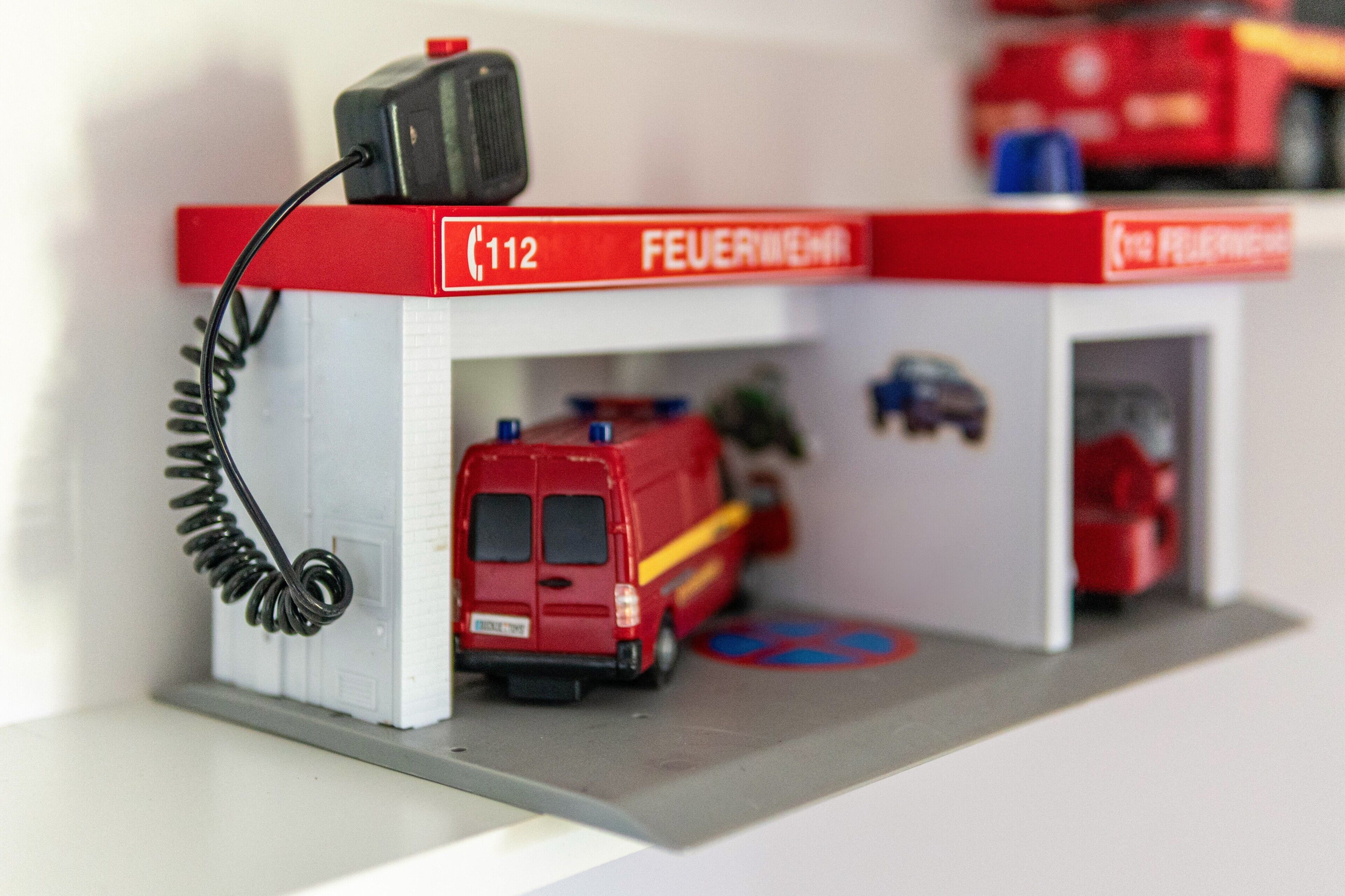 Un modellino di una caserma dei pompieri con un furgone rosso, un camion dei pompieri e un edificio rosso e bianco con la scritta 'FEUERWEHR'.
