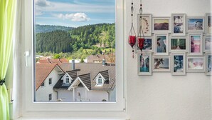 A window with a view of houses and a forest, a curtain, and a wall with framed pictures. - Holiday Apartment 'Fewo Arber up to 10 Guests, Mountain View' with Balcony & Wi-Fi (Frauenau)