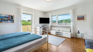 A bedroom with a bed, a TV, a desk, a chair, and a view of the outdoors. - Holiday Apartment 'Fewo Arber up to 10 Guests, Mountain View' with Balcony & Wi-Fi (Frauenau)