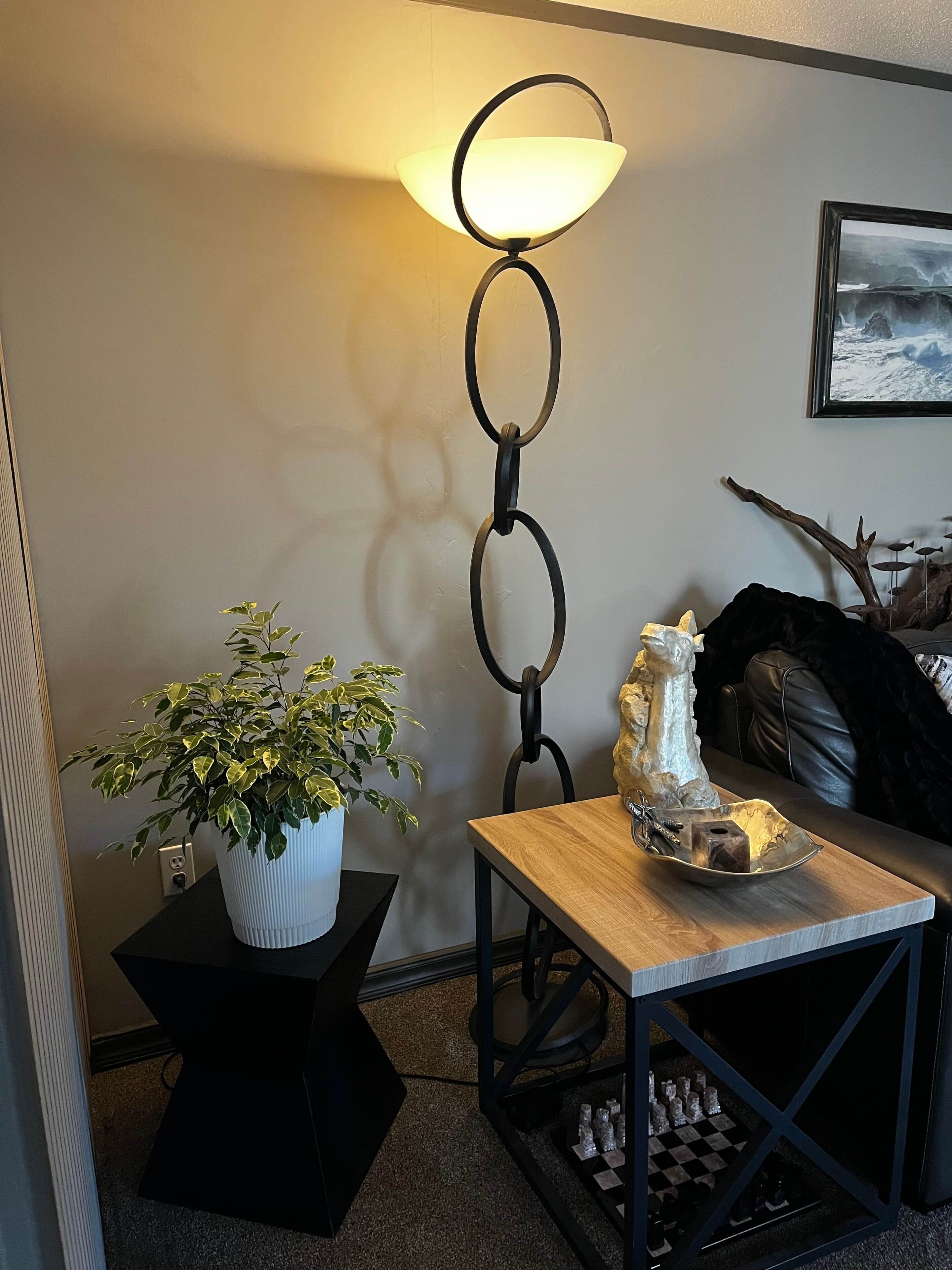 A cozy living room with a potted plant, a table with decorative items, a lamp, and a framed picture on the wall.