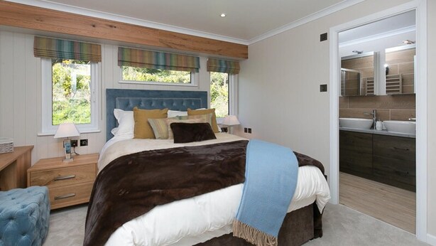 A bedroom with a large bed, a wooden headboard, a nightstand, a blue ottoman, and a view of a bathroom with a sink and mirror. - Quaint Conwy Lodge Retreat (Conwy)