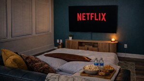 Een slaapkamer met een bed, een televisie waarop 'NETFLIX' wordt weergegeven, een nachtkastje met een kaars en een dienblad met snacks.