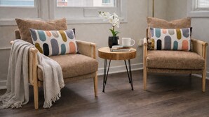 Twee fauteuils met kleurrijke kussens, een klein rond tafeltje met een boek en een kopje, en een vaas met witte bloemen.