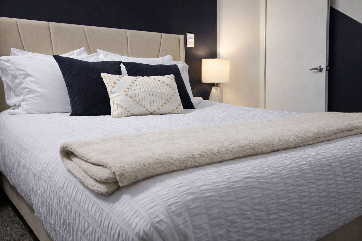 Una cama bien tendida con colcha blanca, un plaid color beige y varios almohadones en blanco, negro y un diseño estampado.