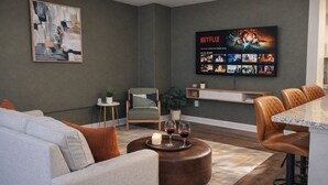 A modern living room with a grey sofa, a coffee table with candles, a TV displaying Netflix content, and a small side table with a plant. - Ste 421 + Free Parking + Skywalk + Downtown 1BR Near Lucas Oil (Indianapolis)