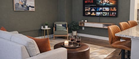 A modern living room with a grey sofa, a coffee table with candles, a TV displaying Netflix content, and a small side table with a plant.