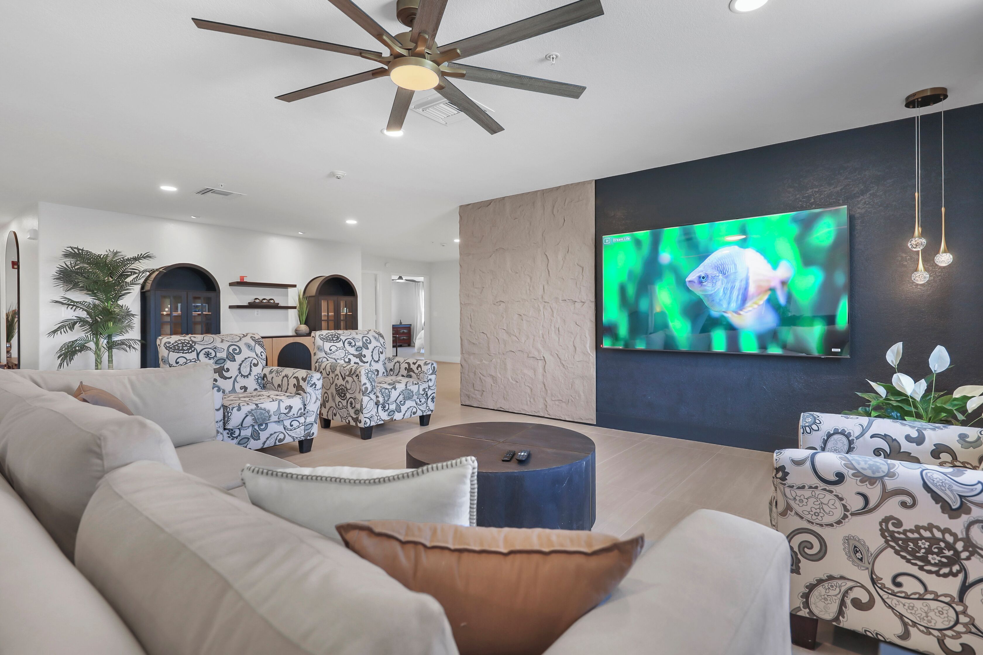 A modern living room with a ceiling fan, a flat-screen TV, patterned sofas, and a round coffee table.