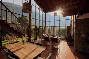 A spacious room with a large wooden table, comfortable seating, and a stone wall. A staircase leads to an outdoor area with a view of mountains. - An impressive house in Peru's Sacred Valley (Calca, Cuzco)