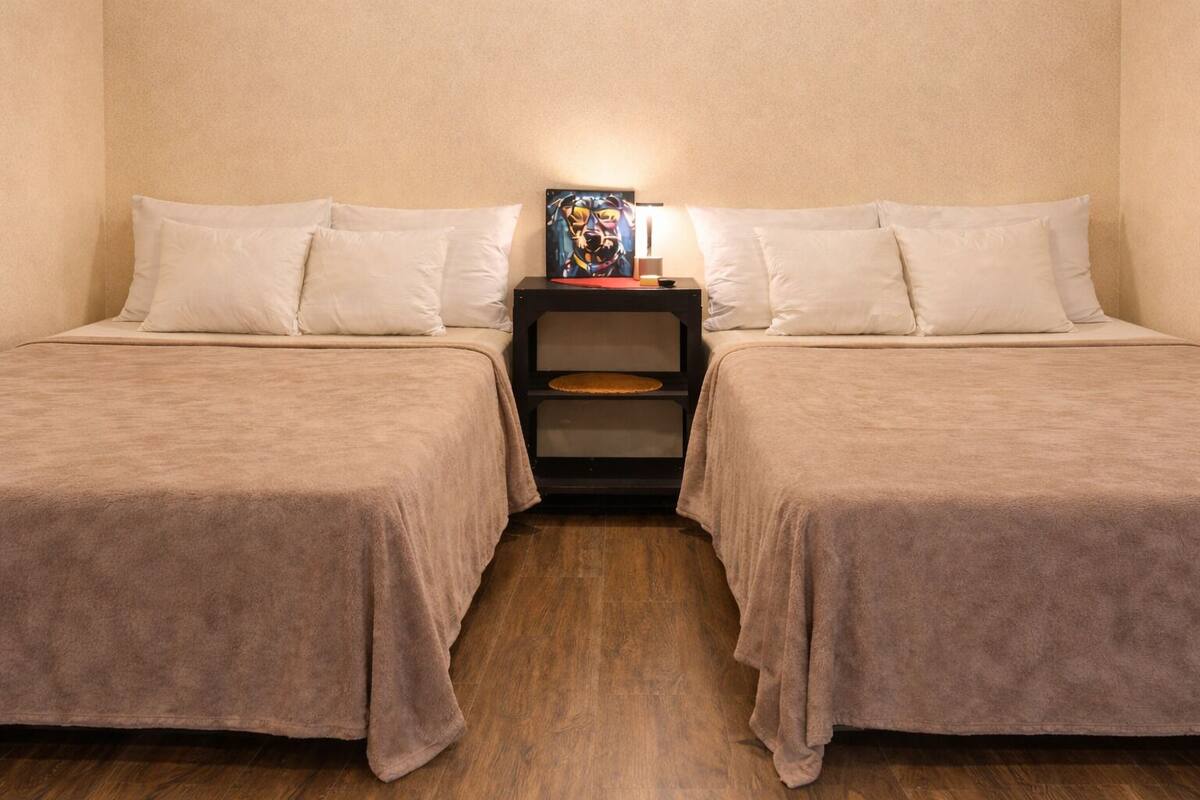 Une chambre avec deux lits, chacun recouvert d’une couvre-lit beige et agrémenté d’oreillers blancs. Une table de chevet se trouve entre les deux lits, sur laquelle reposent une lampe et un tableau encadré. La chambre est dotée d’un plancher en bois.