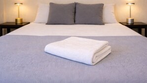 A neatly made bed with a folded white towel, two gray pillows, and a gray bedspread. There are two bedside tables with lamps on either side of the bed. - Los Cipreses Hotel de Montaña (Los Naranjos)