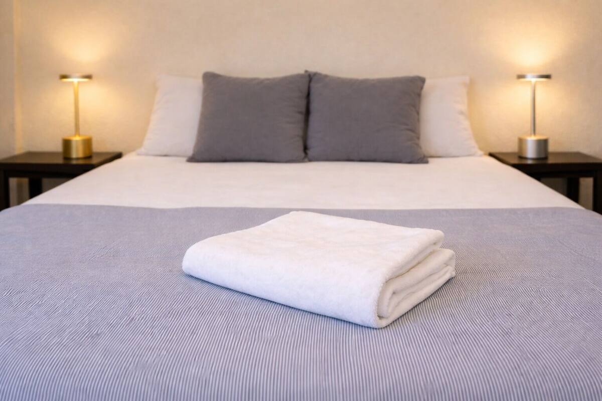 Un lit bien fait, avec une serviette blanche pliée, deux oreillers gris et une couvre-lit grise. De chaque côté du lit, on trouve une table de chevet avec une lampe.