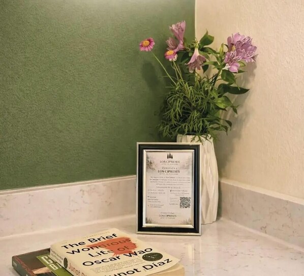 A framed certificate, a book titled 'The Brief Wondrous Life of Oscar Wao', and a potted plant with pink flowers on a hotel counter. - Los Cipreses Hotel de Montaña (Los Naranjos)