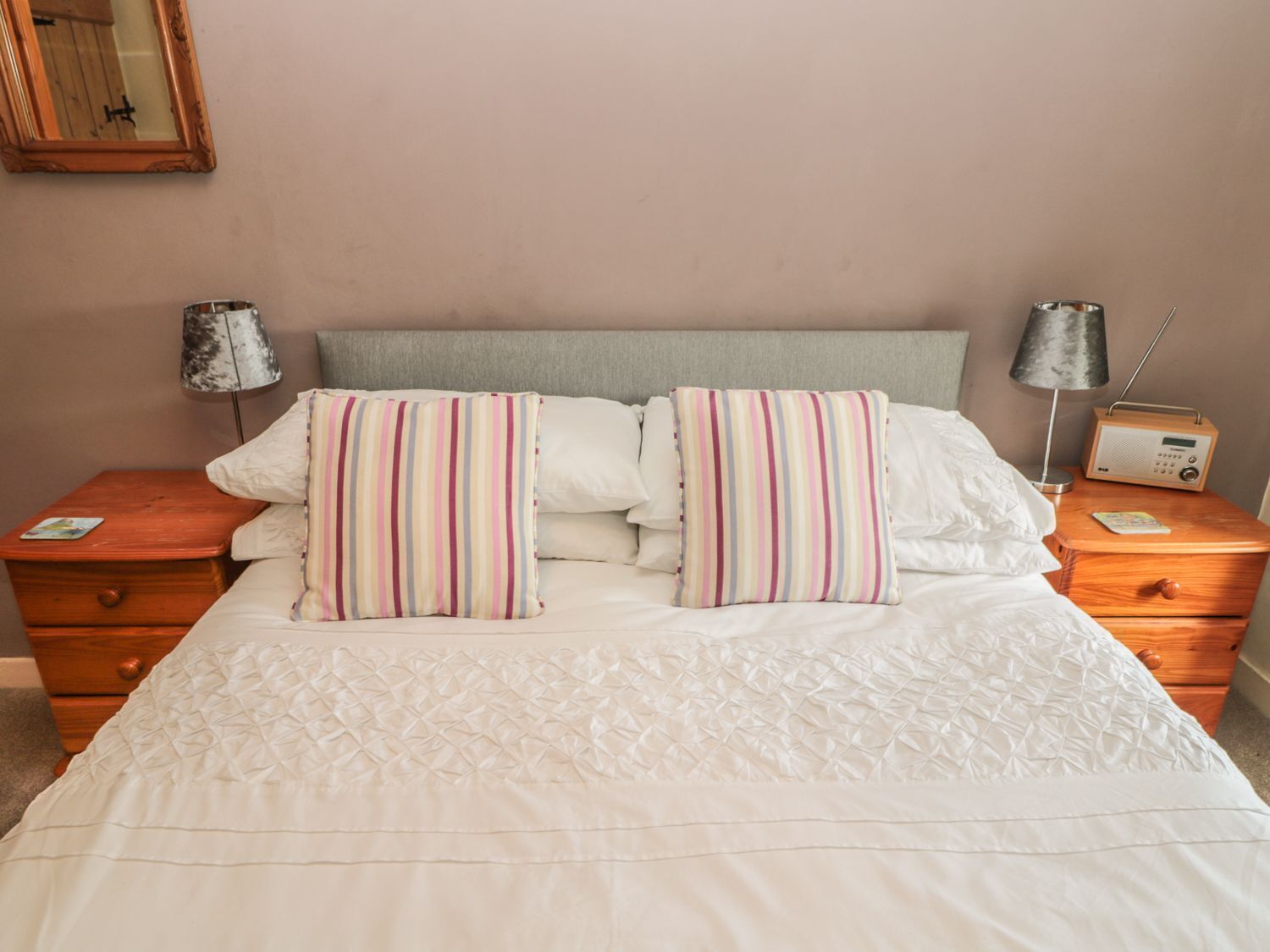 A neatly made bed with striped pillows, a bedside table with a radio, and a mirror on the wall.