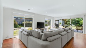 Uma sala de estar ampla com um sofá seccional cinza, uma TV de tela plana em um suporte de madeira e grandes portas de vidro deslizantes que dão para um pátio externo.