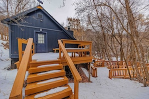 A wooden staircase leading to a blue building with a door and windows, surrounded by snow and trees.