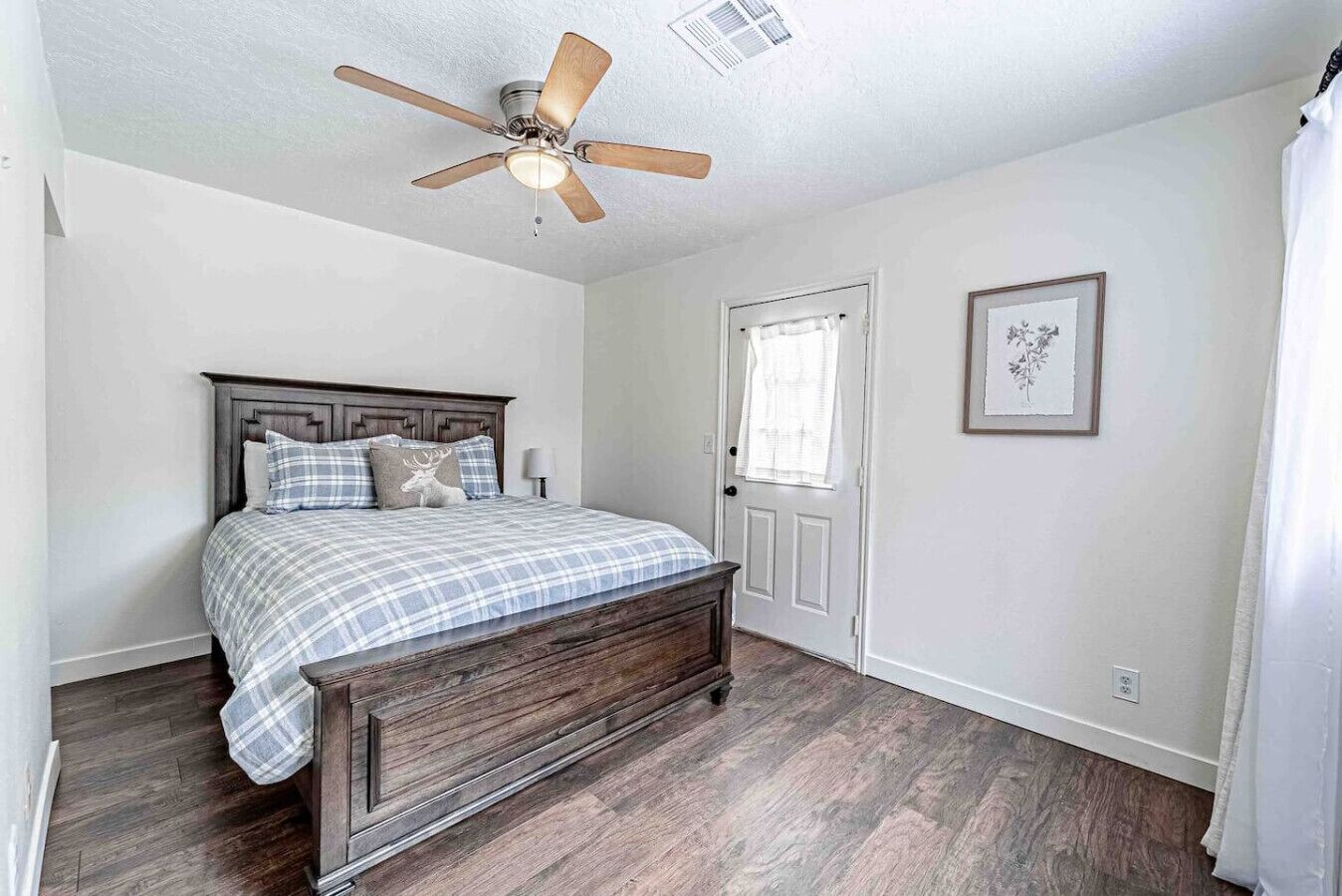 A bedroom with a wooden bed, a ceiling fan, a door with a window, and a framed picture on the wall.