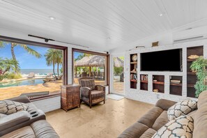 A spacious living room with a large window overlooking a pool and beach, a flat-screen TV mounted on a brick wall, and comfortable seating. - Serenity Bay Estate-FL Keys Best Kept Secret! Beautiful, Quiet, Tropical Pardise (Marathon)