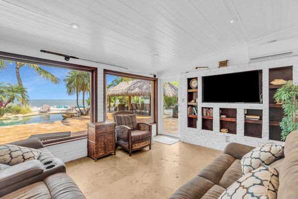 A spacious living room with a large window overlooking a pool and beach, a flat-screen TV mounted on a brick wall, and comfortable seating. - Serenity Bay Estate-FL Keys Best Kept Secret! Beautiful, Quiet, Tropical Pardise (Marathon)
