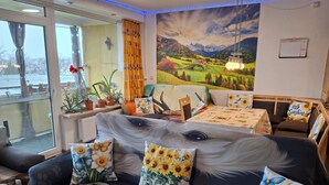 A cozy room with a large window, a sofa with floral cushions, a mural of a landscape, and a table with a floral tablecloth. - App 416 Panoramic Hotel Hohegeiß (Braunlage)