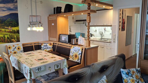 A cozy room with a dining table, benches, and a sofa with floral cushions. A kitchen area is visible in the background with a microwave and sink. - App 416 Panoramic Hotel Hohegeiß (Braunlage)