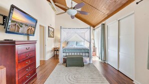 Une chambre à coucher avec un lit sous un dais, un ventilateur de plafond, un téléviseur fixé au mur et une commode en bois.