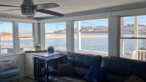 Um quarto com sofá de couro preto, ventilador de teto e janelas amplas com vista para a praia e o mar.