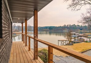 Eine Holzterrasse mit Geländer, die auf einen ruhigen See mit umliegenden Bäumen und einem entfernten Haus blickt.