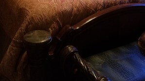 Una cama con colcha estampada y varias almohadas, cabecera de madera, mesita de noche con lámpara y una pequeña mesita auxiliar.