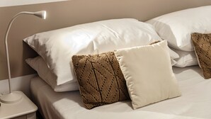 Uma cama bem arrumada com roupa de cama branca e duas almofadas decorativas, uma luminária de cabeceira e uma mesinha de cabeceira branca.